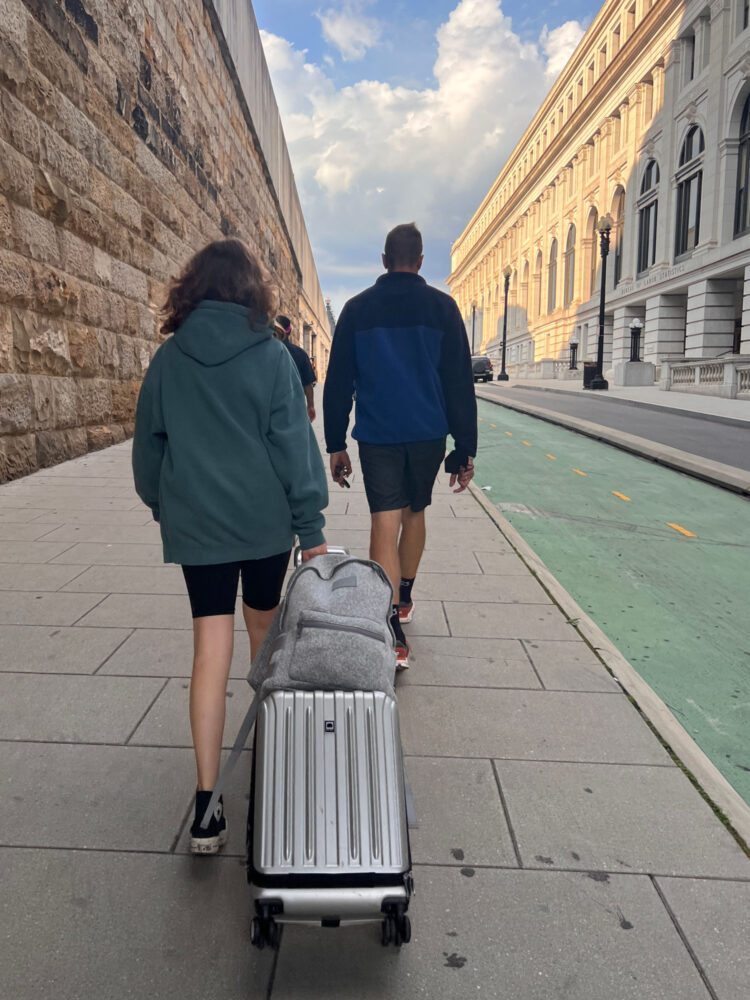 Dagne Dover Dakota Backpack A teen and her father walking into a train station; the teen is pulling a rolling suitcase with a gray Dagne Dover Dakota backpack on top secured by its luggage sleeve. Dagne Dover Dakota backpack review by Wardrobe Oxygen