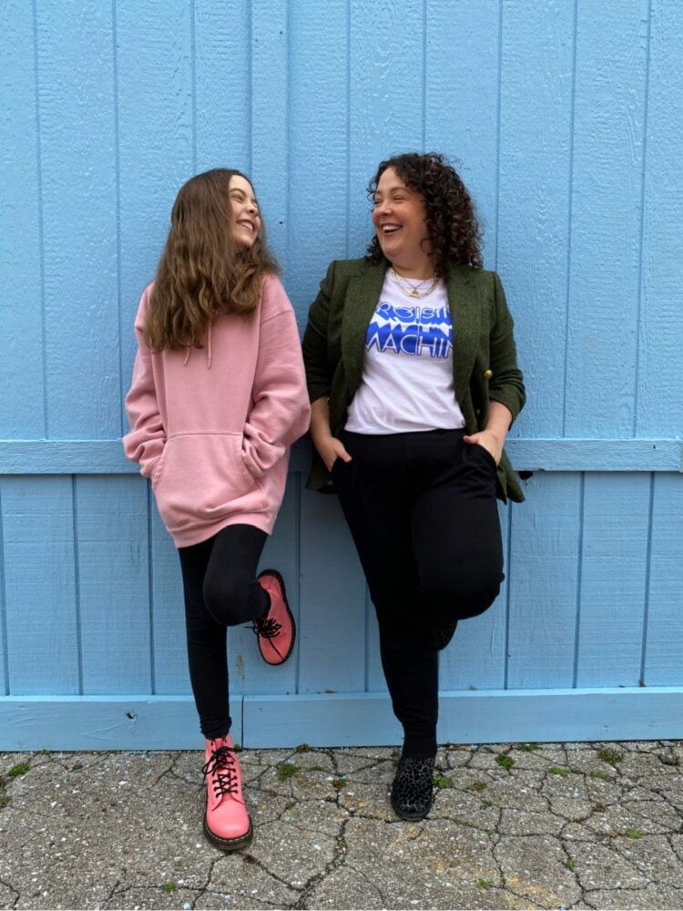 A woman and her tween daughter with their hands in their pockets, laughing with one another while leaning against a wood wall.