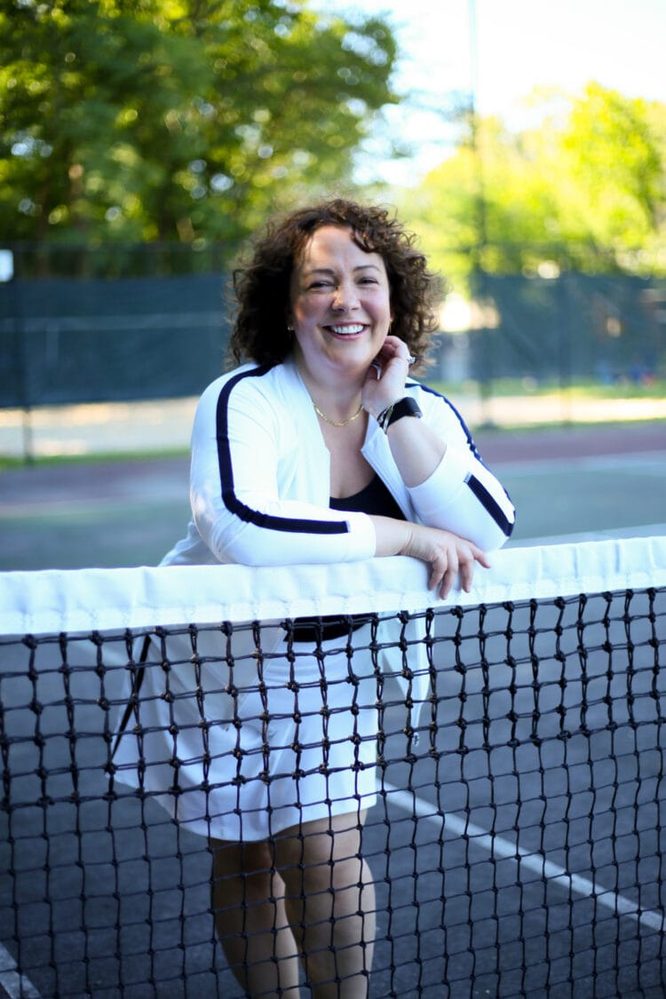 Talbots Tennis Skort and Bomber Jacket 1 Alison in a white knit bomber jacket and matching skort from Talbots. She is leaning against a tennis net smiling at the camera