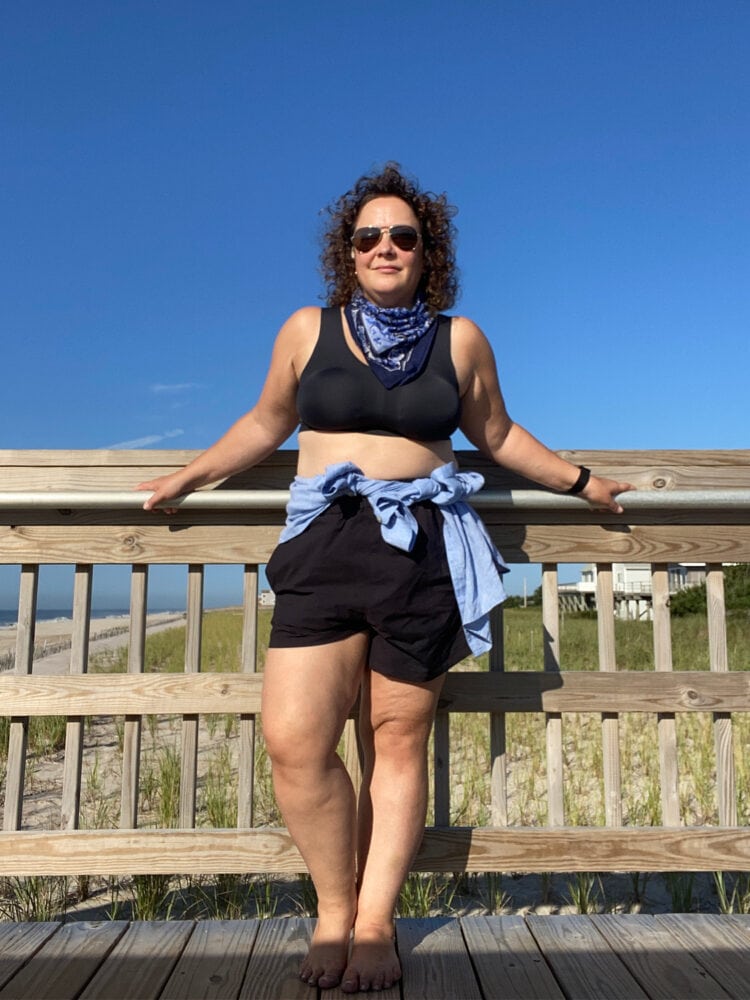 Alison Gary of Wardrobe Oxygen in a black cropped tank, black pull-on shorts, and a blue linen shirt tied around her waist. She is standing on a deck at Fire Island