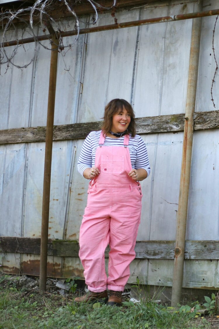 Grapefruit WV Overalls: "She Could Be a Farmer in Those Clothes" 2 Alison of Wardrobe Oxygen in pink Dickies overalls and a black and white striped long sleeved tee. She is standing in front of a metal storage unit with her hands in her pockets smiling away from the camera