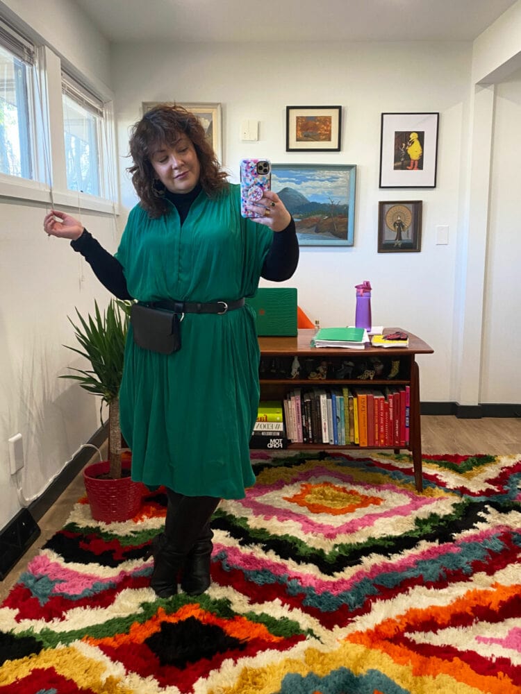 Alison Gary of Wardrobe Oxygen in her office wearing a green ever by X dress over a black turtleneck with a black belt bag and black leather knee high boots