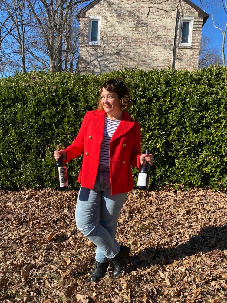 Alison of Wardrobe Oxygen wearing her favorite Breton top under a red wool peacoat. She is standing outside, holding two bottles of wine and smiling at the camera
