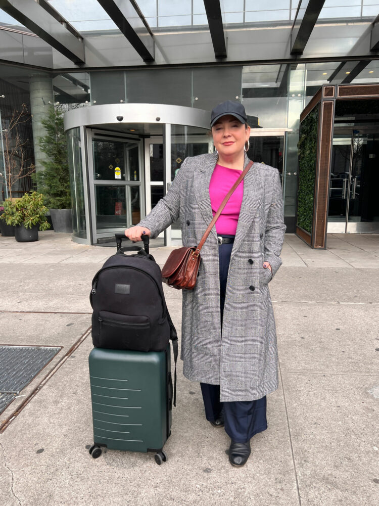 I Tried Quince Luggage: An Honest Quince Suitcase Review 1 Alison Gary of Wardrobe Oxygen outside an NYC hotel holding onto a Quince rolling suitcase that has a Dagne Dover Dakota backpack on top of it.