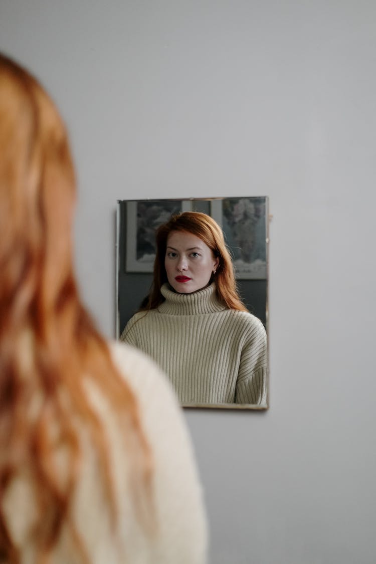 A redhead woman gazes pensively at her reflection in a mirror, wearing a knitted turtleneck indoors.