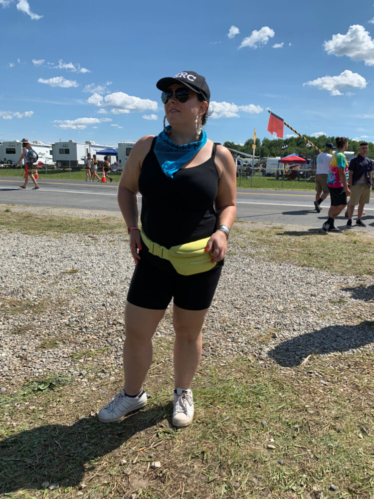 woman in a black tank and bike shorts and a neon belt bag at a music festival. She is over 40 and wearing a baseball cap.