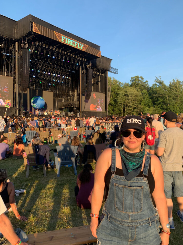 Woman in shortalls and a baseball cap in front of a stage where Vampire Weekend is performing at the 2019 Firefly music festival